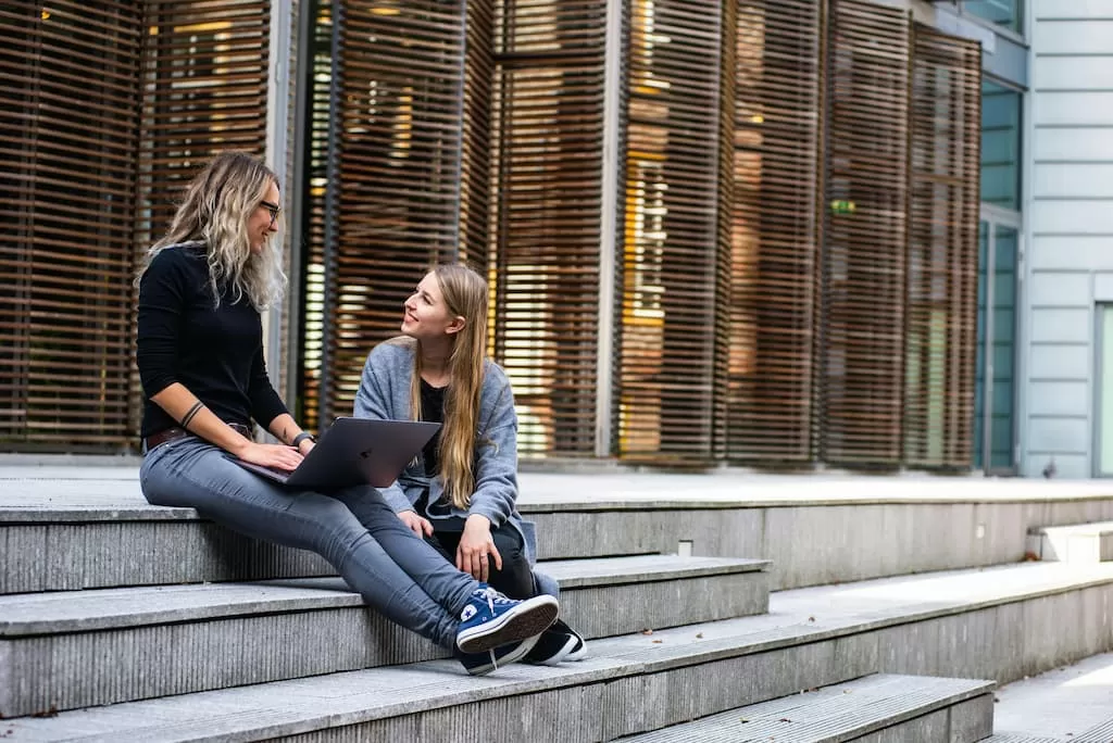 Dos estudiantes hablan sentadas en las escaleras de la entrada de la facultad